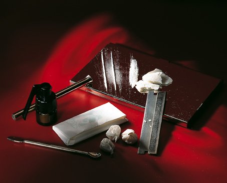 A set of tools on a red background. On the left side of the image there is a black spray bottle with a long handle and a nozzle. Next to the spray bottle there are two small white objects one of which appears to be a piece of paper or a napkin and the other of which is a metal tool with a curved handle. The metal tool is resting on top of a red surface which is covered in a layer of white powder. There are also a few garlic cloves scattered around the tools. The overall color scheme of the background is red and black creating a striking contrast with the white objects.