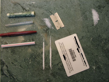 A green countertop with various items scattered on it. On the left side of the countertop there are two red and white pens one with a green cap and the other with a white cap. Next to the pens there is a small piece of paper with a barcode and some text on it which appears to be a label or a bar code. In the center of the image there appears to have a small amount of white powder or sugar scattered around. The items are arranged in a scattered manner with some of them overlapping each other.