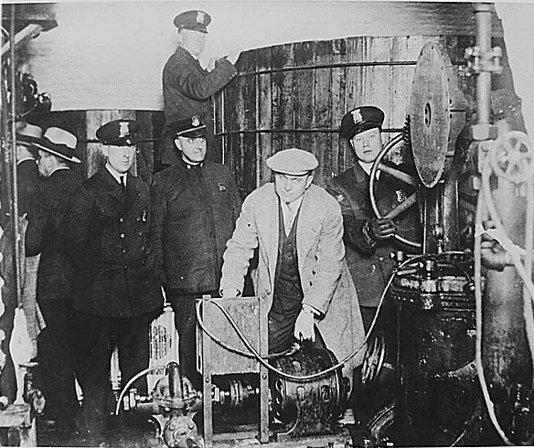 A black and white photograph of a group of men standing in front of a large machine. The machine appears to be a steam engine or boiler with a large cylindrical tank on the right side and several pipes and valves on the left side. The men are wearing suits and hats and one of them is standing on top of the tank while the others are standing around it.<br /><br />In the center of the image there is a man wearing a suit and a hat who is holding a large pipe in his hands. He is standing next to the machine and there are several other men standing around him also wearing hats and jackets. The man in the suit is looking at the machine with a serious expression on his face. The other men in the group are looking at him attentively. The background is blurred but it seems like they are in a room with other machinery and equipment.