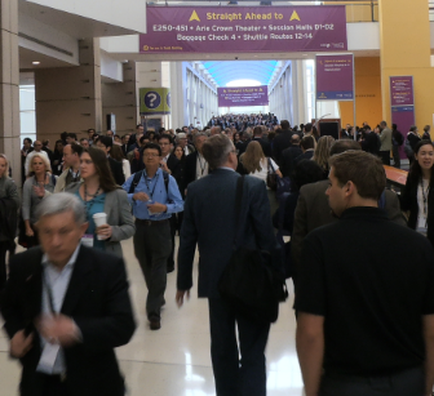 A large group of people walking in a large hall with a high ceiling and columns. The hall appears to be a conference or exhibition hall as there is a banner hanging from the ceiling that reads "Straight Ahead to A". The people in the image are of different ages genders and ethnicity's and they are all dressed in formal business attire. Some of them are carrying bags suggesting that they are attending an event or gathering. The people are walking in different directions and some are looking towards the banner. The overall mood of the image is busy and bustling.