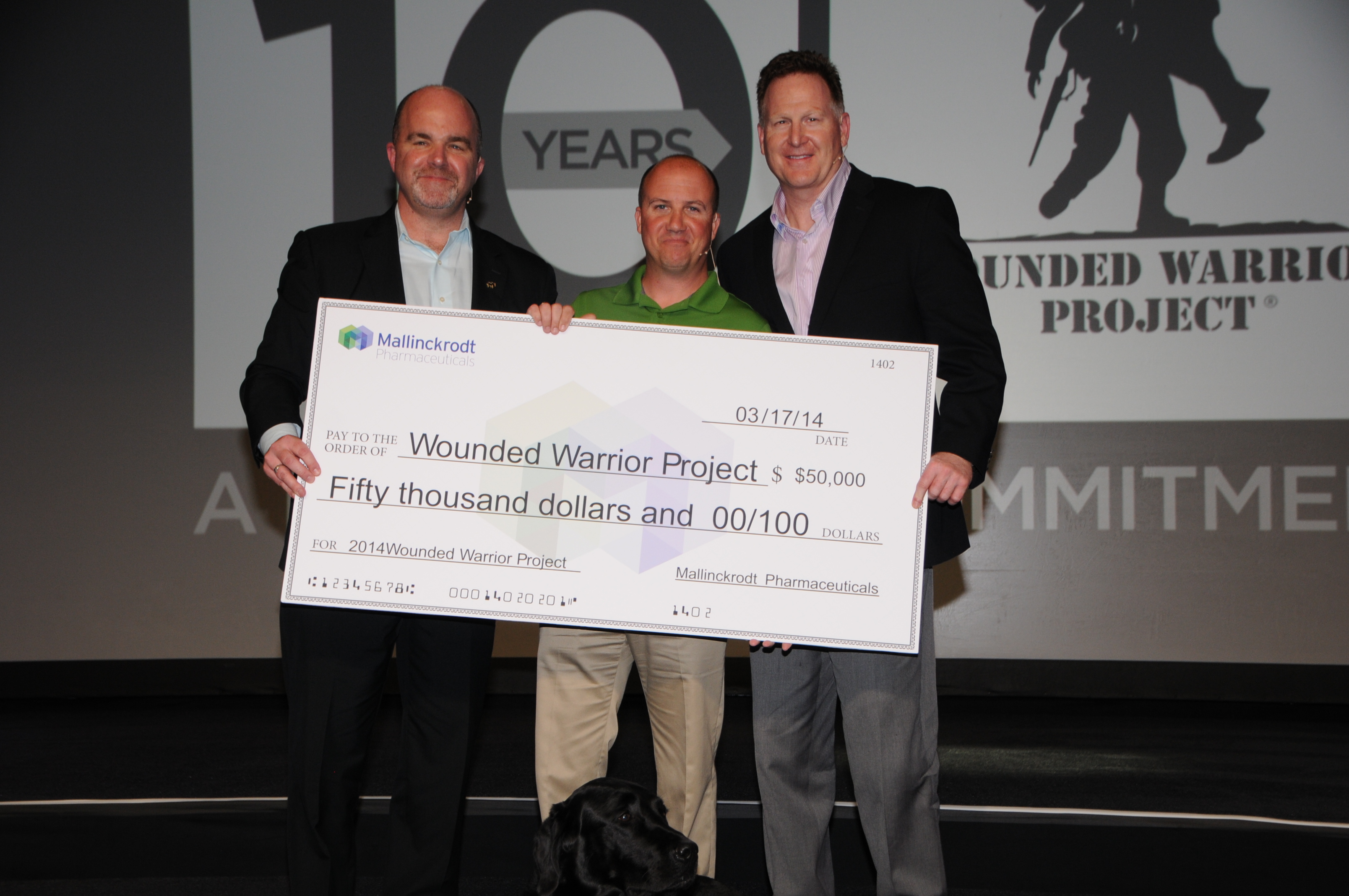 Three men standing on a stage with a large check in front of them. The check is for $50000 and is being presented to the wounded warrior project. The man on the left is holding the check with both hands while the man in the middle is holding it with his hands. All three men are smiling and appear to be proud of the donation. In the background there is a large screen with the logo of the project and the words "Wounded Warrior Project" and "Commitment" on it. A black dog is sitting on the stage next to the check.