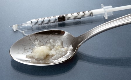 A silver spoon and a syringe next to it. They are resting on a blue grey surface. On the spoon there is a small amount of white substance likely heroin or a another drug. The substance is spilling out of the spoon and onto the tip of the syringe. 