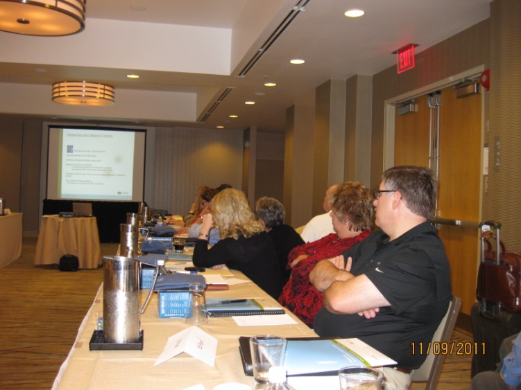 A group of people sitting at a long table in a conference room. The room has a high ceiling with recessed lighting and a projector screen in the background. The people are facing the projector screen and appear to be engaged in a discussion or presentation. The table is covered with a beige tablecloth and there are several water pitchers and glasses on it. On the right side of the image there is a man standing with his arms crossed looking towards the screen. The date "11/09/2011" is visible in the bottom right corner.