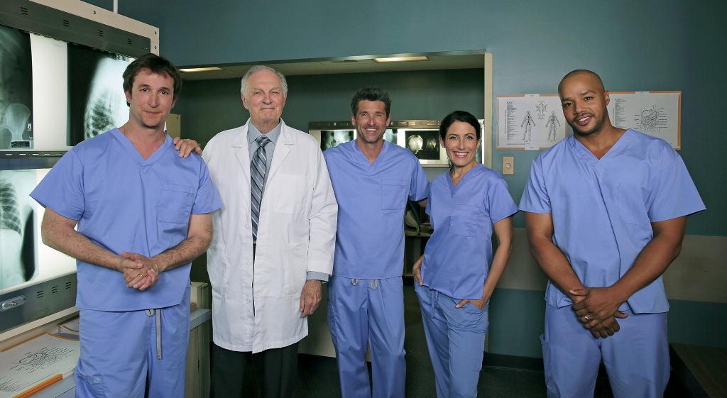 A group of five medical professionals standing in a hospital room. They are all wearing blue scrubs and are standing in front of a large x-ray machine. On the left side of the image there is a man wearing a white lab coat and a tie who appears to be a doctor or a medical professional. Next to him there are two men and two women all of whom are smiling and looking at the camera. The man on the left is standing with his arms crossed while the man in the middle is standing behind him with his hands on his hips. The woman on the right is standing next to him with her hands on her hips and the man at the far right is also smiling. In the background we can see medical equipment and charts on the wall.