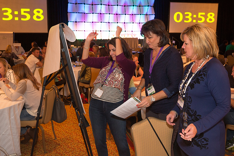 Three women standing in a conference room with a large screen in the background. The screen displays the time as 3:58. The woman in the center is holding a piece of paper and appears to be explaining something to the two women on either side of her. The other two women are looking at the screen and appear to be listening attentively. The room is filled with people sitting at tables and chairs and there is a large window with colorful lights on the wall behind them. The floor is covered with a patterned carpet.