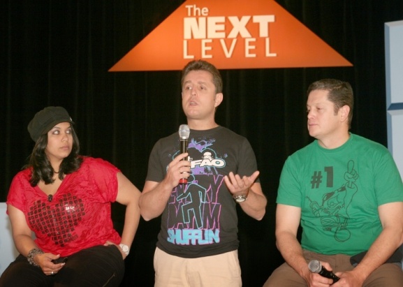 Three people sitting on a stage with a black curtain in the background. On the left side of the image there is a woman wearing a red top and a black beanie. She is sitting next to a man wearing a black t-shirt with the words "The Next Level" written on it. The man in the middle is holding a microphone and appears to be speaking while the man on the right side is listening attentively. All three people are holding microphones and appear to be engaged in a conversation.