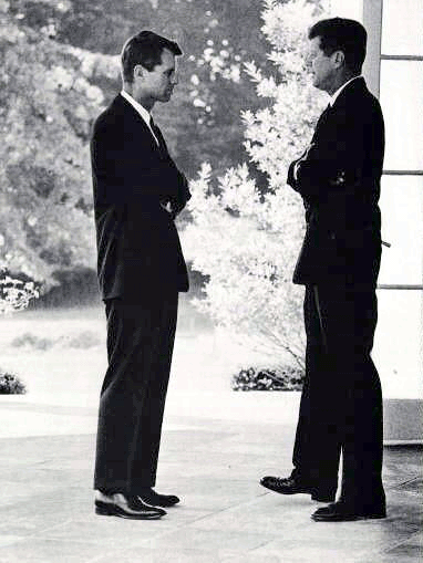 A black and white photograph of two men standing in a room with large windows. They are both wearing suits and appear to be engaged in a conversation. The man on the left is standing with his arms crossed and is looking towards the right side of the image. He has short hair and is wearing a suit and tie. The background shows a garden with trees and bushes. The floor is made of concrete and there is a large window on the right wall. The image appears to be from the early 20th century.