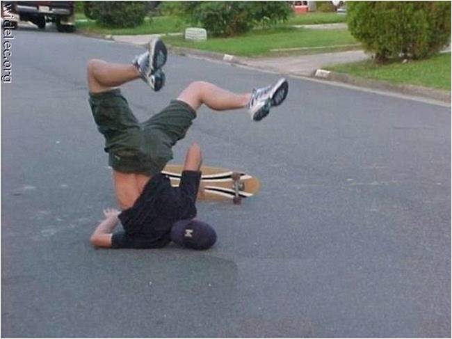 A young man performing a handstand on a skateboard. He is wearing a black t-shirt green shorts and white sneakers. His arms are stretched out to the sides and his legs are bent at the knees. The skateboard is lying on its side on the ground in front of him. The background shows a street with houses and trees. The image appears to be taken on a sunny day.