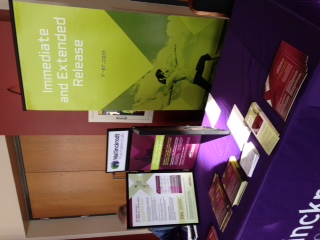 A table with a purple tablecloth and various items on it. On the left side of the table there is a large green banner with the words "Immediate and Extended Release" written in bold white letters. Below the banner there are two smaller banners with the same text and images. The banner on the right side has a picture of a person holding a book and appears to be promoting a product or service. There are also several small pamphlets and brochures scattered around the table. The table is located in a room with a wooden door in the background.