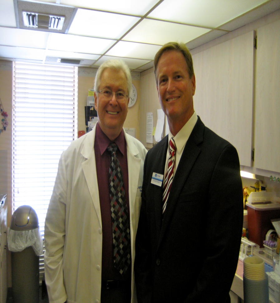 Two men standing side by side in a laboratory. The man on the left is wearing a white lab coat and glasses and he is smiling at the camera. He has short white hair and appears to be in his 60s or 70s. The other man is also wearing a suit and tie. They are both standing in front of a desk with various medical equipment and supplies on it. The background shows a window with blinds and a clock on the wall. Both men appear to be happy and confident.