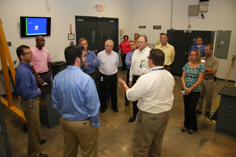 A group of people gathered in a room with a large screen on the wall. There are nine people in the group all of whom appear to be engaged in a conversation. The room has a concrete floor and there are several lockers and cabinets in the background. On the left side of the image there is a ladder leaning against the wall and on the right side there are shelves with various items on them. In the center of the room a man in a white shirt is standing and gesturing with his hands as he speaks to the group. He is wearing a blue shirt and khaki pants. The people around him are of different ages and ethnicities and they are all looking at him attentively.