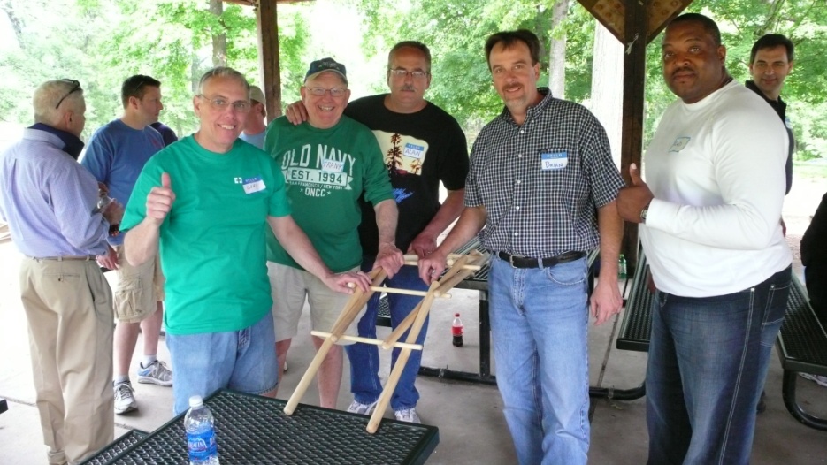 A group of six men standing around a table in a covered area with trees in the background. They are all smiling and appear to be posing for a photo. The man in the center is holding a wooden structure which appears to be a ladder or a ladder and is being held up by one of the men on the left side of the image. On the right side there is another man wearing a green t-shirt with the words "Old Navy" on it. In the background there are other people standing around the table some of them are also smiling and giving thumbs up. There is a water bottle on the table.