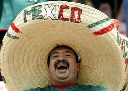 A man wearing a large sombrero with the word "MEXICO" written on it in red and green letters. He is wearing a green and red striped shirt and has a big smile on his face. The background is blurred but it appears to be a stadium with other people in the stands. The man seems to be celebrating or cheering.