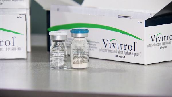 Two vials of Vivitrol a medication used in the pharmaceutical industry. The vials are placed next to a white box with the brand name "Vivitrol" written on it. The box is open and the vials have blue caps. The background is blurred but it appears to be a laboratory setting with other vials and boxes in the background.