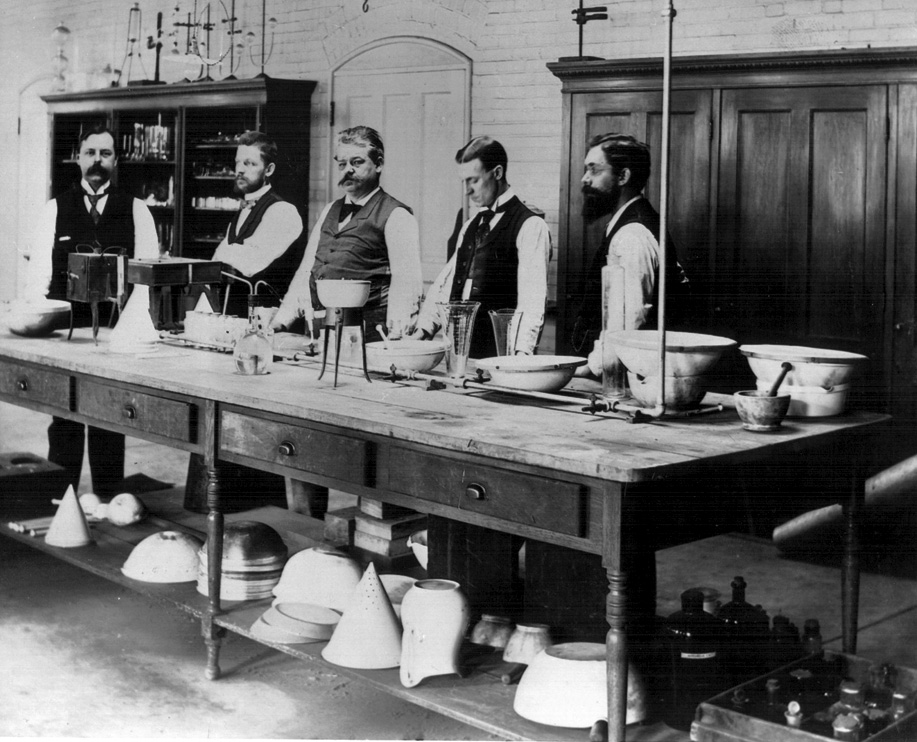 A black and white photograph of a group of six men in a laboratory. They are standing around a long wooden table with various scientific equipment on it. The table is covered with a long tablecloth and there are several bowls plates and other scientific equipment scattered across it. On the left side of the table there is a laboratory bench with a microscope and other laboratory equipment. The men are wearing lab coats and appear to be engaged in a scientific experiment. In the background there are shelves with various tools and equipment including a chandelier hanging from the ceiling. The room appears to be well-lit with natural light coming in from the windows.