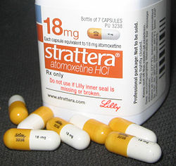 A white container of Strattera Atomoxetine HCI 18mg tablets. The container is cylindrical in shape and has a label on it that reads "18mg" in bold orange letters. Below the label there is a description of the product which states that the tablets are available for purchase at 7 capsules. <br /><br />There are several white and yellow pills scattered around the container. The pills are of different sizes and shapes with some being white and others being yellow. The label also mentions that the product is "Do not use it Lily inner seal is missing or broken."<br /><br />The container is placed on a black surface and there are a few more pills scattered next to it. The background is blurred so the focus is on the container and the pills.