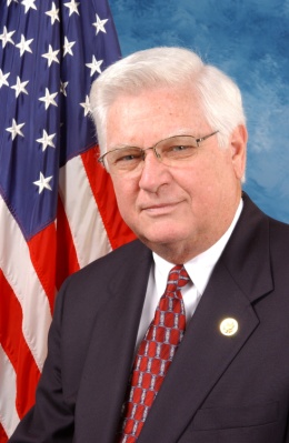 A portrait of a middle-aged man with white hair and glasses. He is wearing a dark suit with a red tie and a gold pin on his lapel. He has a serious expression on his face and is looking directly at the camera. Behind him there is an American flag with red white and blue stripes and stars. The background is a solid blue color.