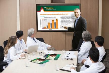 A group of people sitting around a conference table in a meeting room. There are six people in the room all wearing white lab coats and they appear to be engaged in a discussion. The man standing in front of the table is giving a presentation to the group who is smiling and gesturing with his hand as he speaks. On the table there are papers a laptop and a brochure. Behind the man there is a large screen displaying a bar graph and some text. The room has a wooden paneled wall and a carpeted floor.