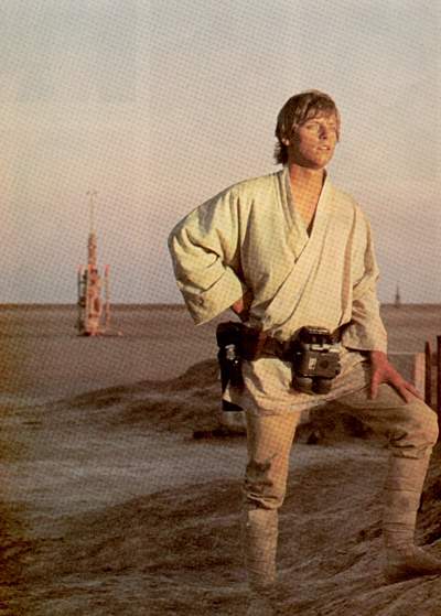 A young man standing on a sandy beach. He is wearing a white robe and has a camera strapped to his waist. He has blonde hair and is looking off to the side with a serious expression on his face. In the background there is a tall tower and a fence. The sky is blue and the overall mood of the image is somber.