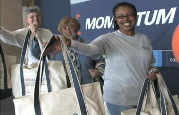 Three women standing in front of a blue background with the word "MOMTUM" written in white letters. They are all smiling and holding large white tote bags with blue handles. The bags appear to be made of a canvas material and have a logo on the front. The women are wearing casual clothes and are standing in a line with one woman on the left one in the middle and one on the right. It seems like they are at an event or gathering.