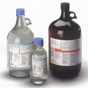 Three bottles of different sizes and shapes. On the left there is a large glass jug with a blue cap and a label on it. The jug has a handle on the top and a spout on the side. The label on the jug is white with black text and has a red and white striped border. Next to it there are two smaller bottles one with a clear glass body and the other with a transparent glass body. All three bottles appear to be made of glass and have a cylindrical shape. The background is plain white.