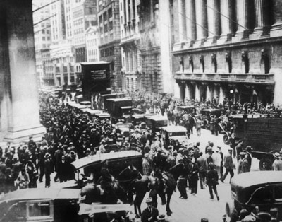 A black and white photograph of a busy street in New York City. The street is filled with a large crowd of people some of whom are standing on the sidewalk and some are walking on the street. There are several cars and trucks on the road and a large group of people can be seen in the background. The buildings on either side of the street are tall and ornate with columns and arches. The sky is overcast and the overall mood of the image is somber. The photograph appears to be from the early 20th century.