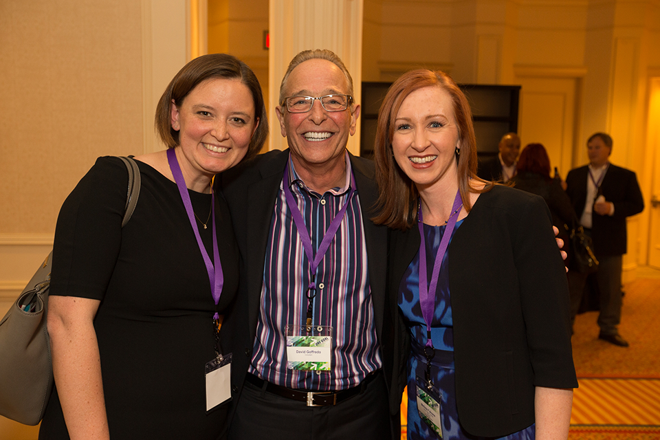 Three people standing in a large room with a high ceiling and chandeliers. The person in the middle is an older man with glasses and a striped shirt. He is smiling and posing for the photo with two women on either side of him. All three are wearing purple lanyards around their necks. The woman on the left is wearing a black dress and carrying a gray purse. The man on the right is also wearing a blue dress and a black blazer. They are all smiling and appear to be happy. In the background there are other people in the room and a carpeted floor.