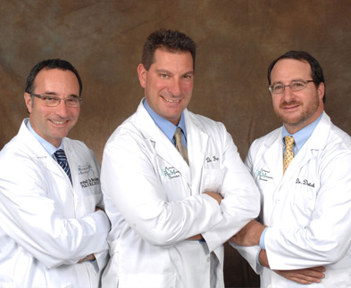 Three men in white lab coats standing side by side with their arms crossed. They are all smiling and appear to be in a professional setting. The man on the left is wearing a blue collared shirt and tie the man in the middle has short dark hair and is wearing glasses and the man next to him has a beard and mustache. All three men have name tags on their lab coats indicating that they are medical professionals. The background is a plain beige color.