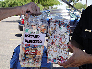 Two large glass jars filled with colorful pills and capsules. The jars are being held by a person wearing a blue shirt who appears to be a police officer. The jar on the left has a label that reads "Expanded Medicines" and the jar in the middle has the same label as the one on the right. The background shows a parking lot with cars and trees.