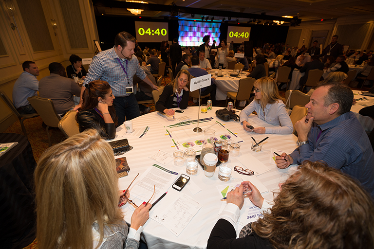 A group of people sitting around a round table in a conference room. The table is covered with a white tablecloth and has various items on it including papers pens cups and other office supplies. There are several people sitting at the table some of them are engaged in conversation while others are looking at the screen in the background. A man in a blue shirt is standing at the front of the table and appears to be giving a presentation. The room is filled with other people and there are large screens on the walls displaying the time. The overall atmosphere of the room is professional and organized.