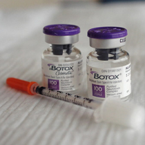 Two small vials with purple caps and white labels. The vials are labeled "BOTOX" and "100". Next to the vials there is a syringe with an orange handle. The syringe is lying on a white surface. The background is blurred but it appears to be a desk or table.