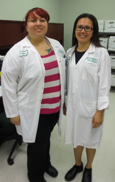 Two women standing side by side in a laboratory. They are both wearing white lab coats with green labels on them. The woman on the left has red hair and is wearing a pink and white striped shirt and black pants. She has a tattoo on her left arm and is smiling at the camera. The other woman is wearing glasses and is also smiling. In the background there are shelves with medical supplies and a desk with a computer monitor. Both women appear to be happy and confident.