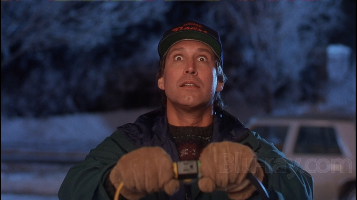 A still from a movie or TV show. It shows a man wearing a green jacket and a red baseball cap with a logo on it. He is holding a pair of gloves in his hands and appears to be looking up at the sky with a surprised or shocked expression on his face. The background is blurred but it seems to be a snowy landscape with trees and a car visible in the distance. The overall mood of the image is one of excitement or surprise.