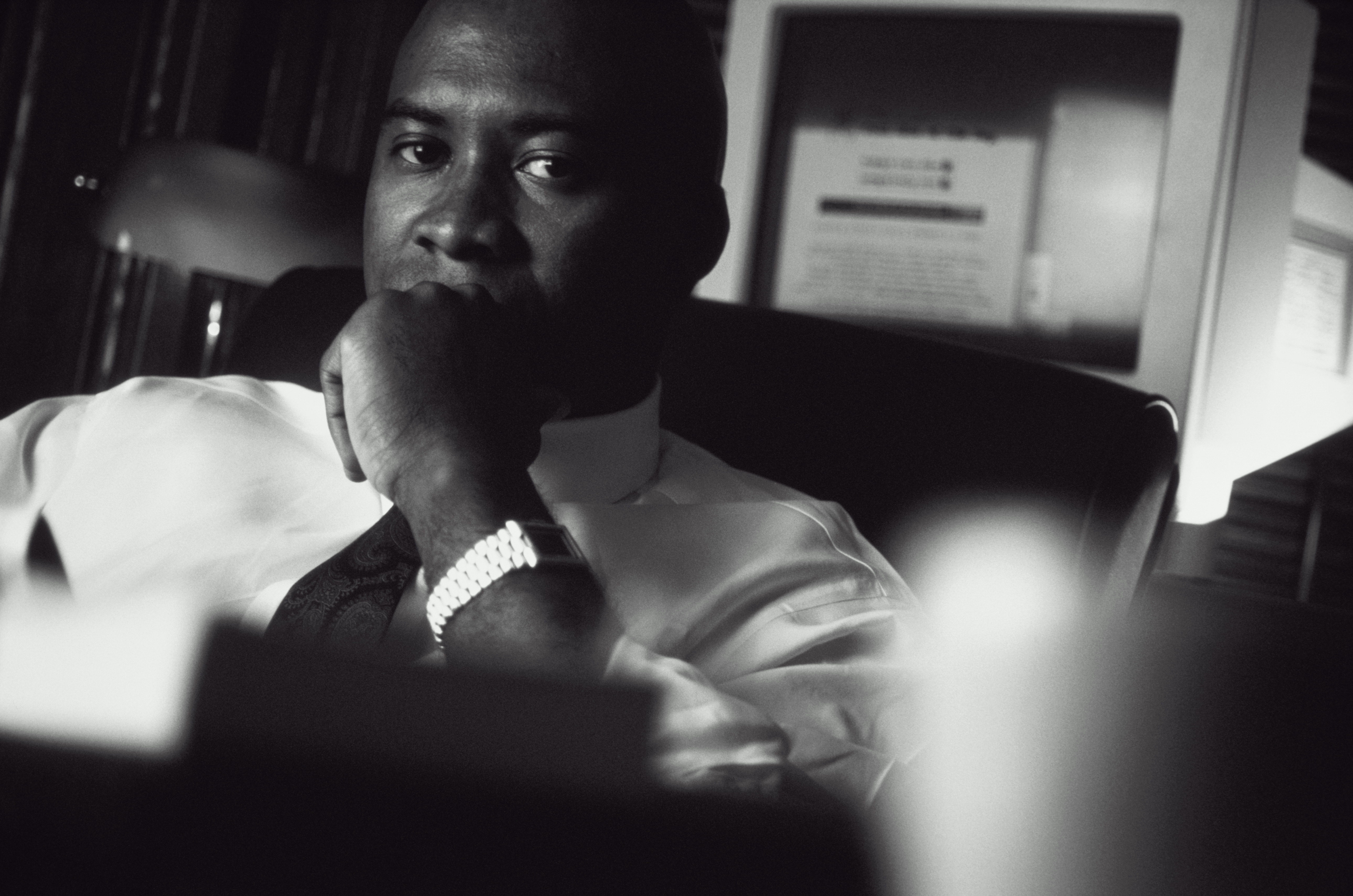 A black and white photograph of a man sitting in an office chair. He is wearing a white shirt and a tie and has a watch on his left wrist. The man is looking off to the side with a thoughtful expression on his face. He has his hand resting on his chin as if deep in thought. The background is blurred but it appears to be a desk with a computer monitor and other office supplies. The lighting is dim creating a moody atmosphere.