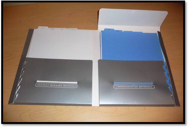 Two folders one in grey and the other in blue. The folders are open revealing the contents inside. The grey folder on the left has a label that reads "District Manager Materials" and the blue folder has the words "Representative Materials" written on it. Both folders appear to be made of metal and have a rectangular shape. The blue folder is slightly larger than the grey folder and has a flap on top. The folder is resting on a wooden surface.