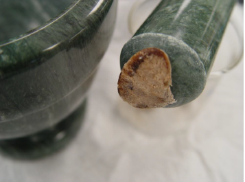 A close-up of a green mortar and pestle. The mortar is made of ceramic and has a round base with a handle on one side. On the other side of the mortar there is a small piece of bread with a brownish-brown crust on top. The bread appears to be freshly baked and is resting on the handle. The background is blurred but it seems to be a white surface.