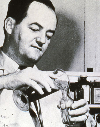 A black and white photograph of a man in a laboratory setting. He is holding a small glass flask with a funnel attached to it and appears to be pouring something into it. The man is wearing a lab coat and has a serious expression on his face. In the background there are shelves with various bottles and jars on them. The photograph appears to have been taken in the early 20th century.