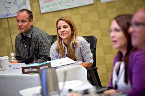 A group of three people sitting at a table in a conference room. The table is covered with a white tablecloth and has a nameplate a water bottle and some papers on it. The person in the center is a young woman with long brown hair and is smiling at the camera. She is wearing a striped shirt and appears to be engaged in a conversation with the other two people. On the right side of the image there is a man and a woman who are also sitting at the table. The background is a yellow wall with a map and a sign that reads "Welcome to the conference".