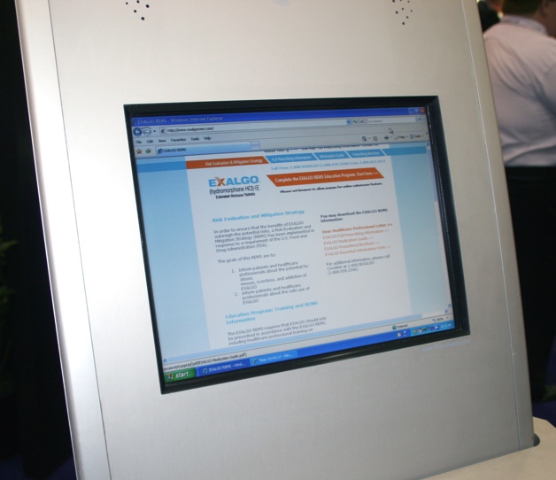 A computer monitor mounted on a white wall. The monitor is displaying a webpage with the word "EXALGO" written in orange and blue text. The webpage appears to be a website or application for a company called EXALGO. The background of the webpage is white and there are a few people visible in the background. The image appears to have been taken at an exhibition or trade show.