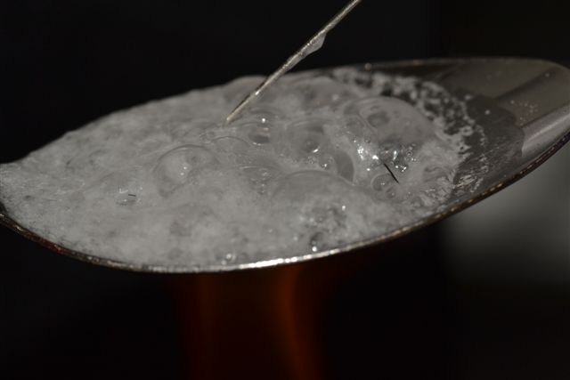 A close-up of a spoon with a white substance on it likely heroin or another drug. The substance is bubbling as though it was just heated. A tip of a syringe is in the substance.  The background is dark making the spoon the focal point of the image.