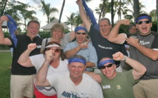 A group of people posing for a photo in a park or garden. There are nine people in the photo six men and three women all wearing blue bandanas on their heads. They are all smiling and appear to be happy and excited. The man in the center is wearing a white t-shirt with the words "Air Force" written on it. The other people around him are also smiling and cheering. In the background there are palm trees and a building. The sky is blue and the weather appears to be sunny and warm.