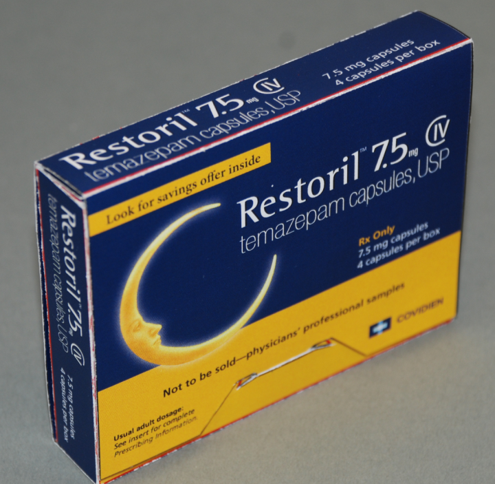 A photograph of a blue and yellow box of Restoril 7.5 mg capsules. The box is rectangular in shape and has a white background. The front of the box has the brand name "Restoril" written in bold white letters at the top followed by the product name "7.5mg Capsules" in smaller white letters. Below that there is an image of a crescent moon with the text "Look for savings offer inside" and "Temazepam capsules USP" in yellow letters. <br /><br />The box also has a warning label that reads "Not to be sold-physicians' professional samples" in black letters. The label also mentions that the capsules are not available for sale. There is also a small illustration of a syringe on the bottom right corner of the label.