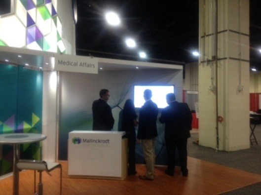 A medical affairs booth at an exhibition. The booth is white with a colorful geometric design on the left side and a sign that reads "Medical Affairs". There is a small table in front of the booth and a few people standing around it. On the right side of the image there is a large screen displaying information about the booth. The people in the image appear to be engaged in a conversation. The background is dark suggesting that the exhibition is taking place at night.