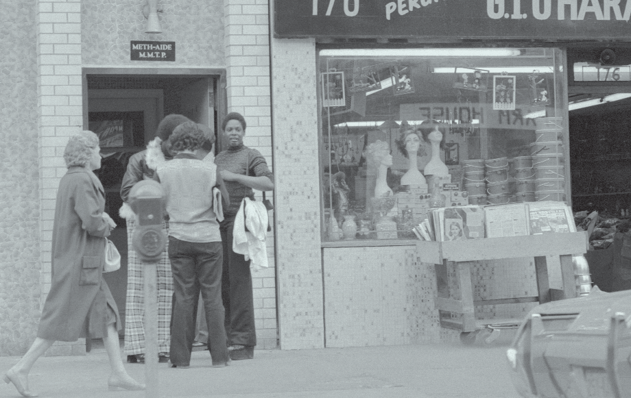 A black and white photograph of a group of people standing outside of a store named "170 Peruvian U.I.U.Hara". The store is located on a street corner with a brick building on the left side and a car parked on the right side. The store has a large window display with mannequins and other items for sale. There are three people in the foreground two women and one man standing in front of the store. The woman on the far left is wearing a long coat and carrying a bag while the woman in the middle is holding a jacket. The man on the top left is standing behind the woman and the man on top right is standing next to her. All three people appear to be looking at the items on display.