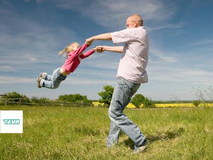 A father and daughter playing in a field. The father is holding the daughter's hand and they are both smiling. The daughter is in mid-air with her arms stretched out to the sides and her legs bent at the knees. She is wearing a pink shirt and blue jeans. The field is green and there is a wooden fence in the background. The sky is blue and there are trees in the distance. The image appears to be taken during the day.