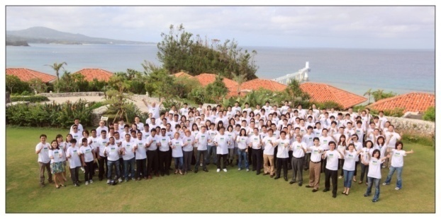 A large group of people standing on a grassy hill overlooking the ocean. They are all wearing white t-shirts and are arranged in a circle forming a heart shape. In the background there are houses with red roofs and a view of the ocean and mountains. The sky is blue and the weather appears to be sunny and warm. The group seems to be posing for a group photo or group photo.