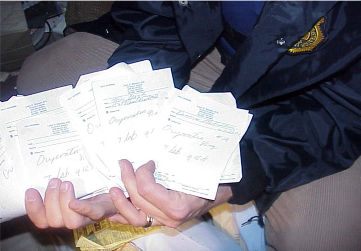 A person's hands holding a bunch of papers. The papers appear to be receipts or bills with some of them having text written on them. The person is wearing a black jacket with a yellow emblem on the left side of their chest. The background is blurred but it appears to be an indoor setting with a couch and other items visible.