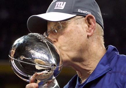 An elderly man wearing a blue New York Giants cap and a blue jacket. He is holding a large silver trophy in his right hand and is kissing it tenderly. The background is blurred but it appears to be a football stadium with spectators in the stands. The man is likely a coach or a member of the New England Patriots football team.