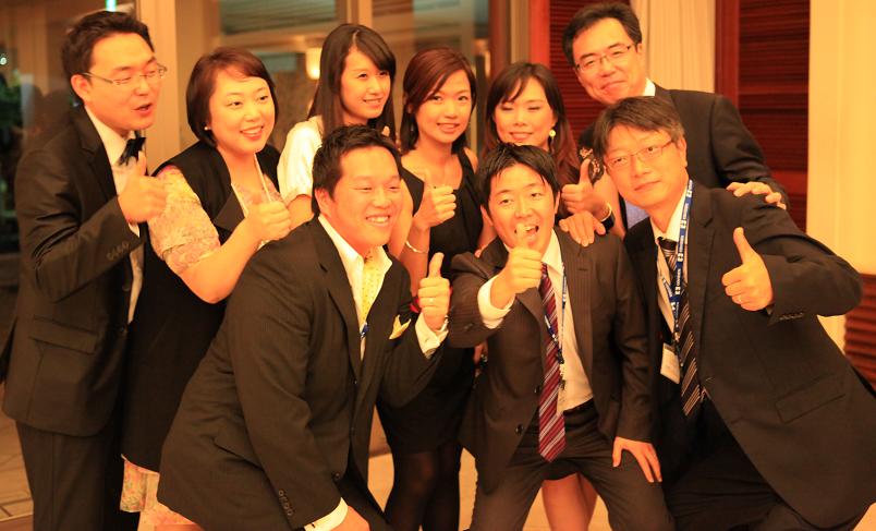 A group of nine people posing together in a room with a large window in the background. They are all dressed in formal attire with the men wearing black suits and the women wearing black dresses. The group is smiling and giving a thumbs up indicating that they are happy and excited. The overall mood of the image is celebratory and happy.