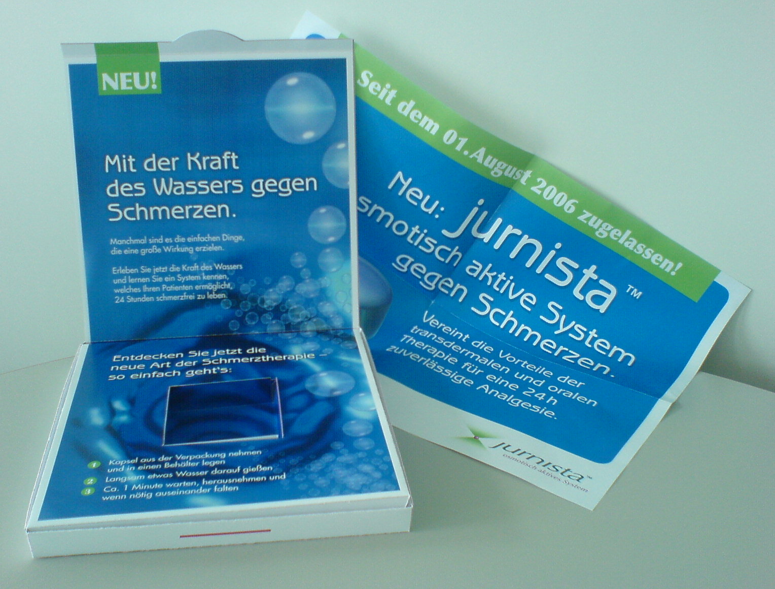 Two brochures one in blue and the other in green side by side. The blue brochure on the left has a white background with a blue cover that reads "Mit der Kraft des Wasser gegen Schmerzen" which translates to "With the Kraft of the Water System" in English. The cover also has an image of a blue water droplet and the text "Jurnista" in white. On the right side of the brochure there is a green banner with the same text. The banner also has the same blue background and white text. Both brochures appear to be new and unused.